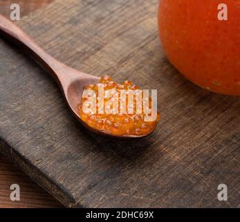 Red caviar in a wooden brown spoon on the table, behind a full jar of caviar Stock Photo