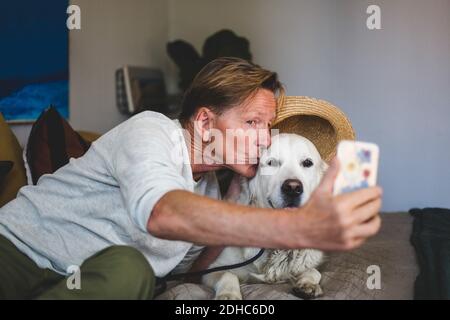 Senior man kissing dog while taking selfie on smart phone in bedroom at home Stock Photo