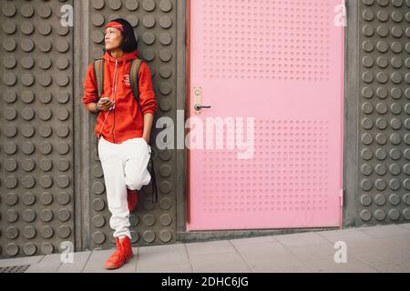 Thoughtful young man looking away while holding mobile phone against wall Stock Photo