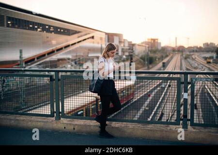 Young woman using mobile phone while leaning on railing at bridge in city during sunset Stock Photo