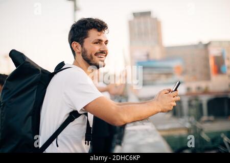 Side view of smiling young man looking away while holding mobile phone at bridge Stock Photo