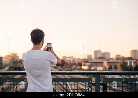 Rear view of young man photographing city through mobile phone while standing on bridge against clear sky Stock Photo