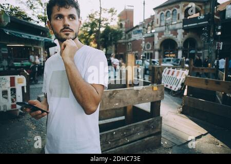 Thoughtful young man holding mobile phone while standing on street in city Stock Photo