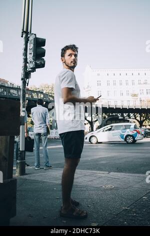 Side view of young man looking away while standing on sidewalk in city Stock Photo