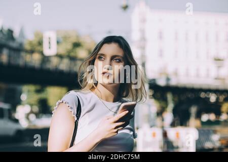 Thoughtful young woman holding mobile phone while standing on street in city Stock Photo