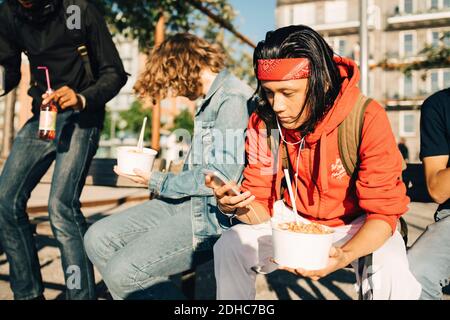 Young man using mobile phone while having take out food by friends in city Stock Photo