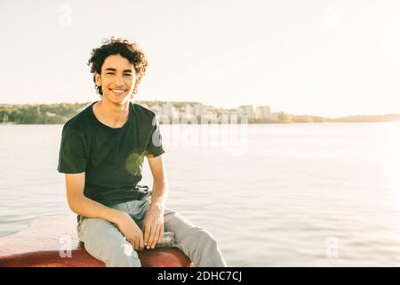 Portrait of smiling teenage boy sitting on bollard by river during sunny day Stock Photo