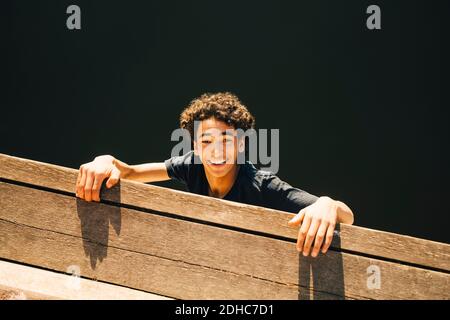 High angle portrait of cheerful teenage boy hanging from pier against river Stock Photo