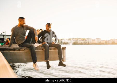 Full length of friends talking while sitting on pier over river against clear sky Stock Photo