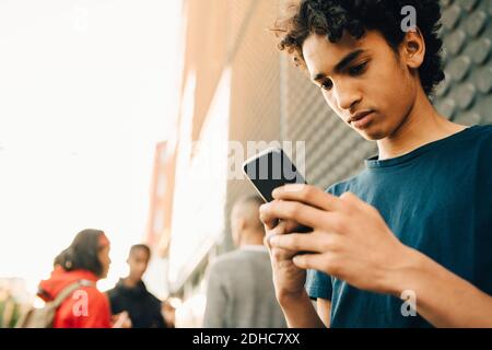 Teenage boy using mobile phone while friends standing in background on city street Stock Photo