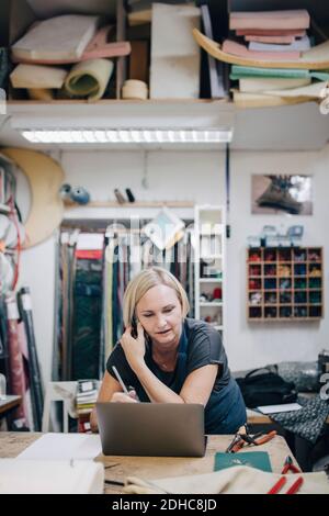 Mature female upholstery worker looking at laptop while talking on phone in workshop Stock Photo