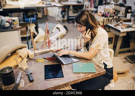 Female upholstery worker talking on phone while using laptop at workbench in workshop Stock Photo
