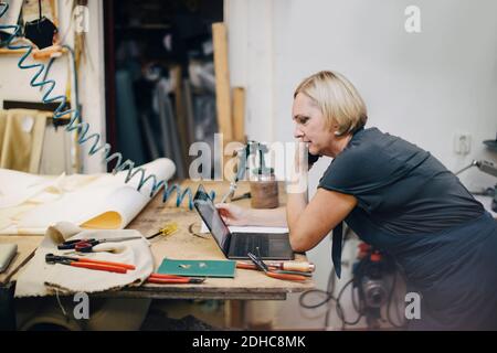 Side view of mature female upholstery worker looking at laptop while talking on phone in workshop Stock Photo