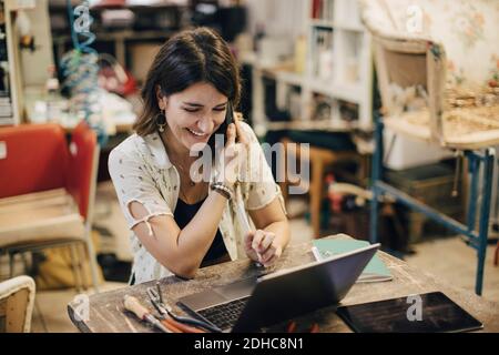 Smiling female upholstery worker using laptop while talking on phone at workbench in workshop Stock Photo