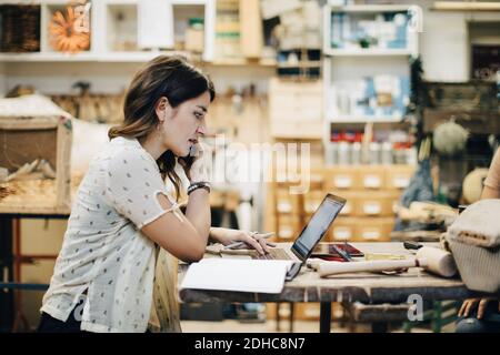 Side view of female upholstery worker using laptop while talking on phone at workbench in workshop Stock Photo