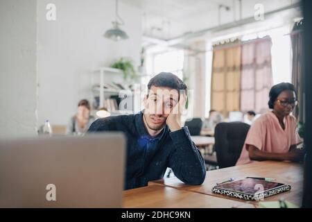 Tired businessman looking at laptop while sitting at desk in creative office Stock Photo