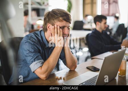 Tired businessman looking at laptop while sitting at desk in creative office Stock Photo