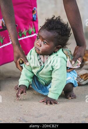 Ranohira, Madagascar - April 29, 2019: Unknown little Malagasy kid, barefoot, crawling on dusty footpath, mother helping her getting up. People in Mad Stock Photo