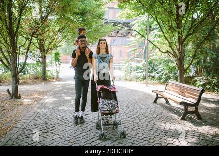 Young man carrying daughter on shoulders while woman pushing baby stroller at footpath Stock Photo