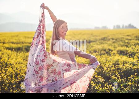 Young long hair woman dancing in yellow flowers field, scarf she is holding moving by wind, afternoon sun backlight background Stock Photo