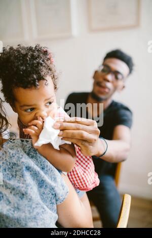 Young man wiping mouth of daughter being held by woman at home Stock Photo