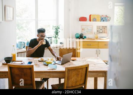 Young man drinking juice while using laptop at table in house Stock Photo