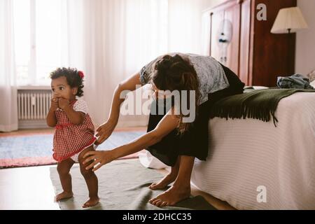 Mid adult woman adjusting diaper of daughter while sitting on bed at home Stock Photo