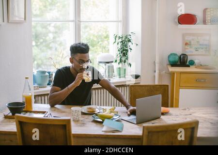 Young man drinking juice while looking at laptop at table in house Stock Photo