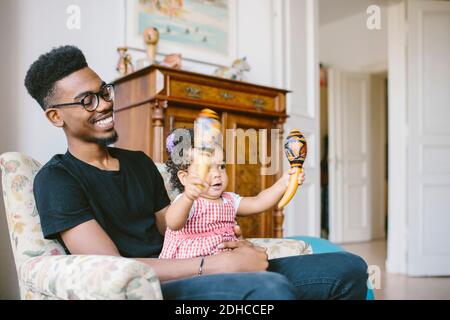Baby girl holding maracas while sitting with happy father on armchair at home