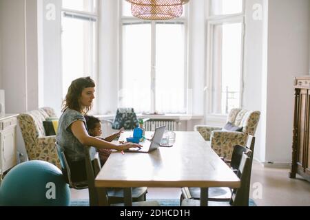 Mother using laptop while sitting with baby girl at table in house Stock Photo