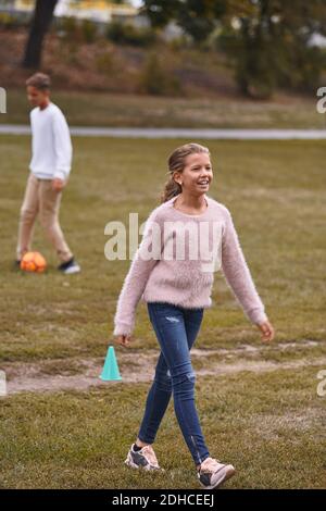 Smiling sister walking on grassy field while brother playing soccer in background at park Stock Photo