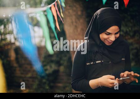 Smiling young woman in hijab using mobile phone at backyard Stock Photo
