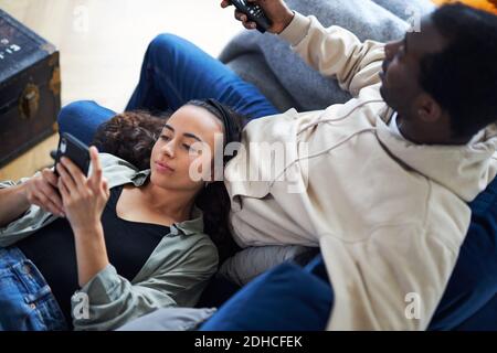 Overhead shot of young mixed ethnicity couple relaxing on sofa at home with man watching tv whilst woman lies across lap and checks social media on mo Stock Photo