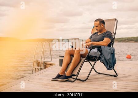 Full length of mature man using laptop while sitting on deck chair at jetty Stock Photo