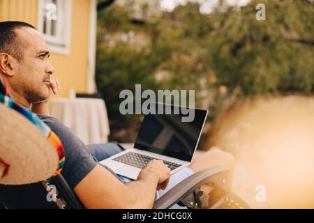 Thoughtful man using laptop while sitting at holiday villa during summer Stock Photo