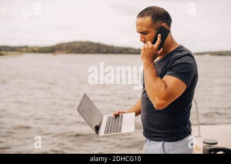 Mature man talking on mobile phone while using laptop by lake Stock Photo