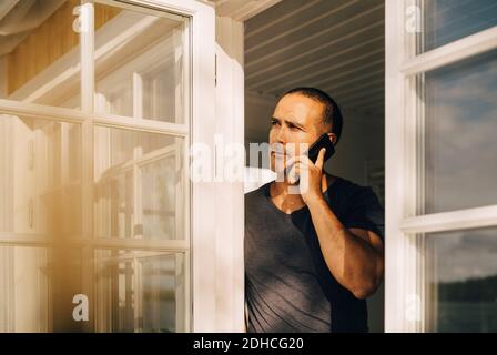 Thoughtful man talking on smart phone while standing at window Stock Photo