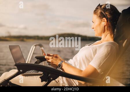 Thoughtful woman with laptop using mobile phone against lake Stock Photo