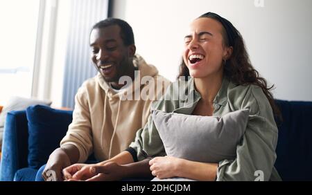Laughing young mixed ethnicity couple laughing whilst sitting on sofa at home watching or streaming comedy show on tv together Stock Photo