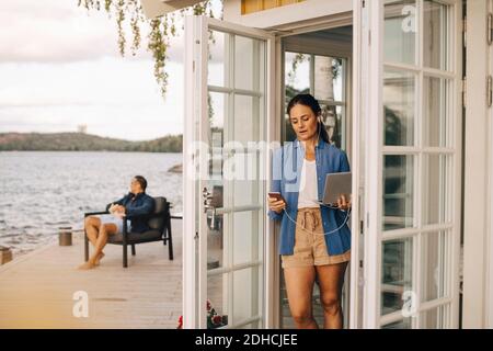 Mature woman using mobile phone and laptop with man in background Stock Photo