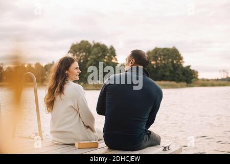 Smiling couple talking while sitting on jetty by lake against sky Stock Photo