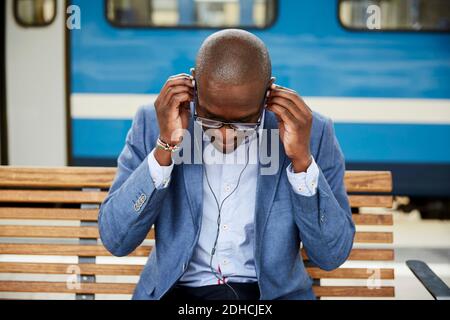 Businessman adjusting in-ear headphones while sitting at railroad station Stock Photo