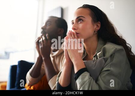 Excited and tense young mixed ethnicity couple sitting on sofa at home watching or streaming sports event on tv together Stock Photo
