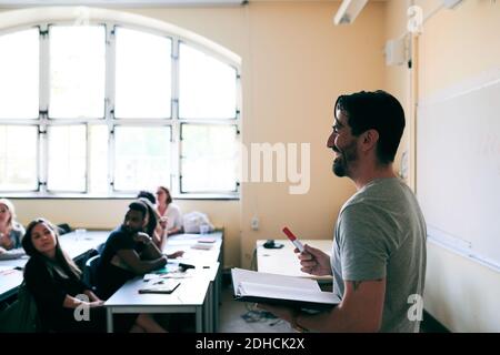 Happy teacher holding book and felt tip pen while teaching language to students Stock Photo