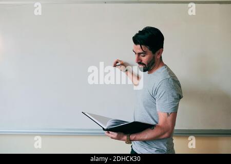 Confident male teacher holding book while writing on whiteboard Stock Photo