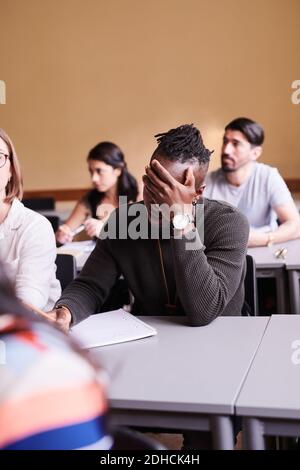Bored young man writing while sitting at desk in language school Stock Photo