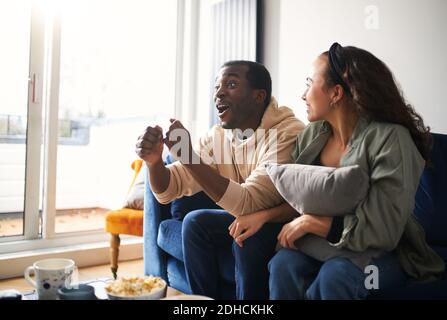 Excited and tense young mixed ethnicity couple son sofa at home celebrating victory of their team whilst watching or streaming sports event on tv toge Stock Photo