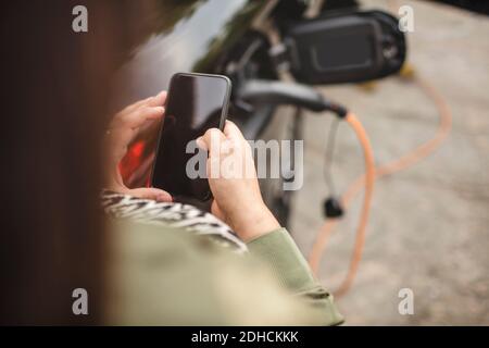 Midsection of woman using smart phone while standing by charging electric car Stock Photo