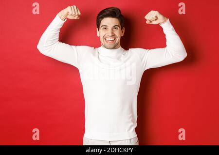 Portrait of smiling handsome man in white sweater, flexing biceps and bragging with strength, show-off strong muscles after work Stock Photo