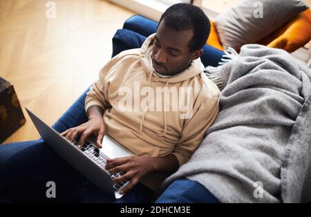 Overhead view of young mixed ethnicity man lying on sofa at home using laptop computer to work from home or shop online Stock Photo